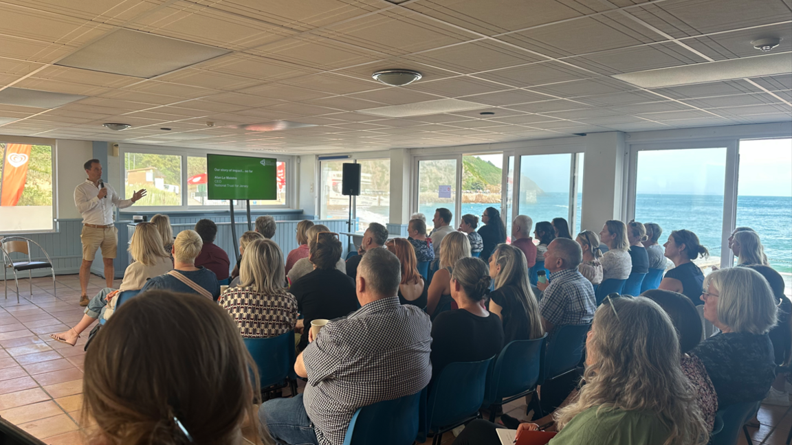 A group of people sat in blue chairs listening to a speaker at the front of the room. There is a green presentation to the right of the speaker at the front, who is holding a microphone. The room has large windows with a sea view.