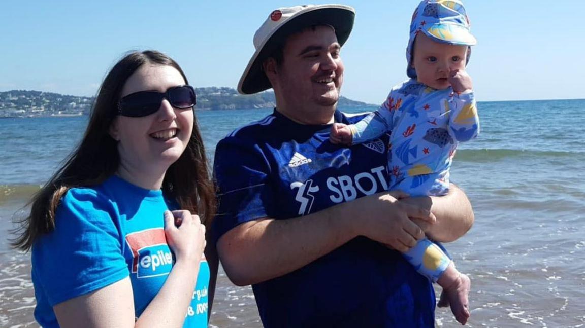 A man and a woman stand together at the seaside smiling. The sea is behind them and the man is holding up a small baby.