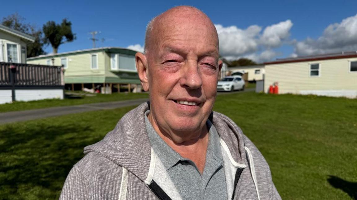 John standing on a patch of grass with caravans behind. He is bald and clean shaven and is wearing a grey zip-up sweater and a grey polo shirt. Blue skies can be be seen in the background. 