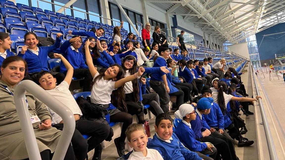 Pupils, some with arms outstretched, look at the camera, sitting on blue seats near a pool to the right. Many are wearing blue.