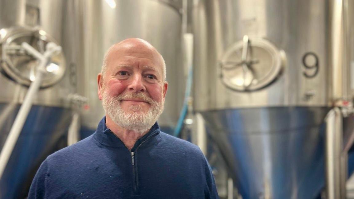 Alan Collyer is wearing a blue quarter-zip sweater and standing in front of a silver vat of beer