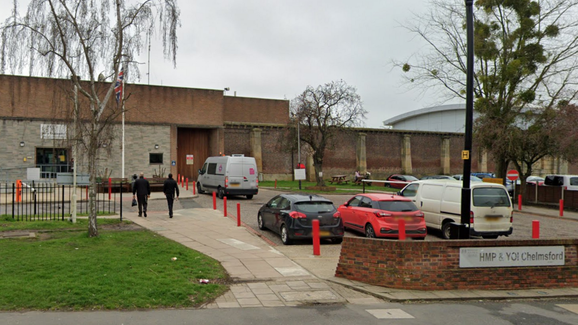 HMP Chelmsford seen from the street. It has large brick walls and has cars parked outside. There is a silver sign that reads "HMP & YOI Chelmsford".