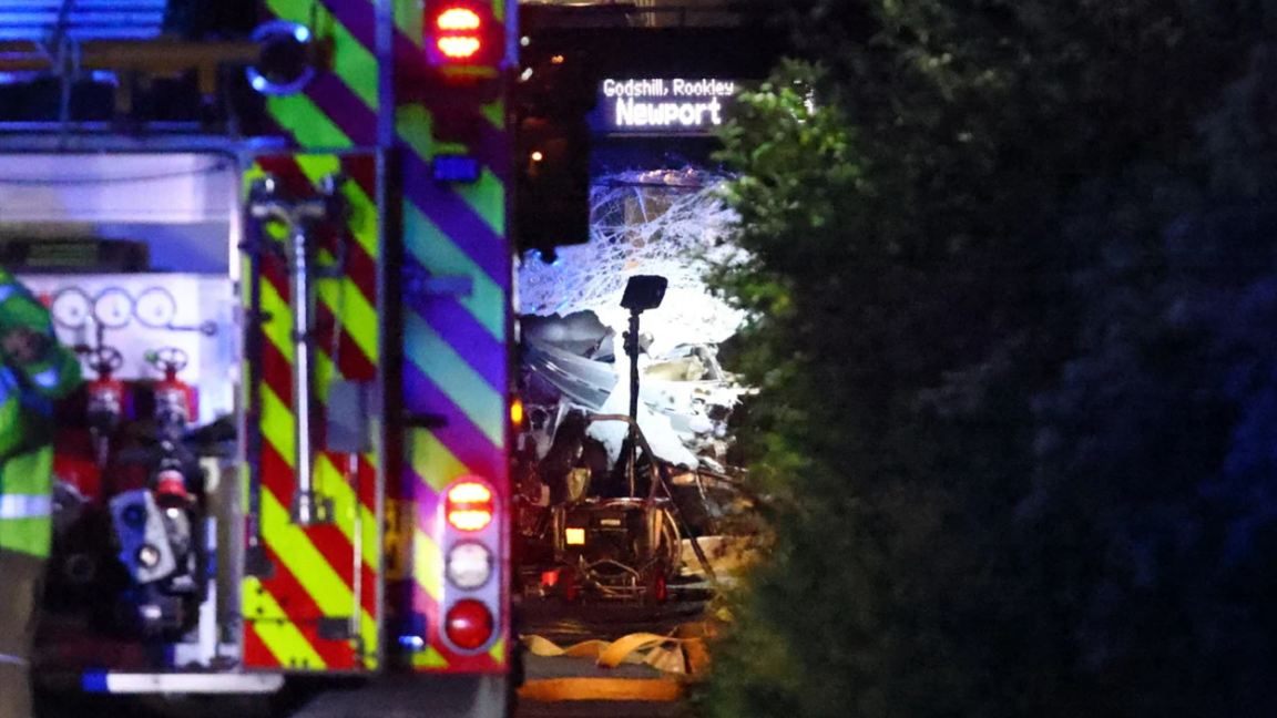 The windscreen of a bus which is smashed. In the foreground, the back of an emergency services vehicle can be seen.