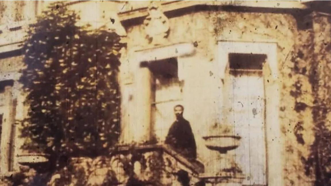 A sepia image of a man standing on steps outside a big house.