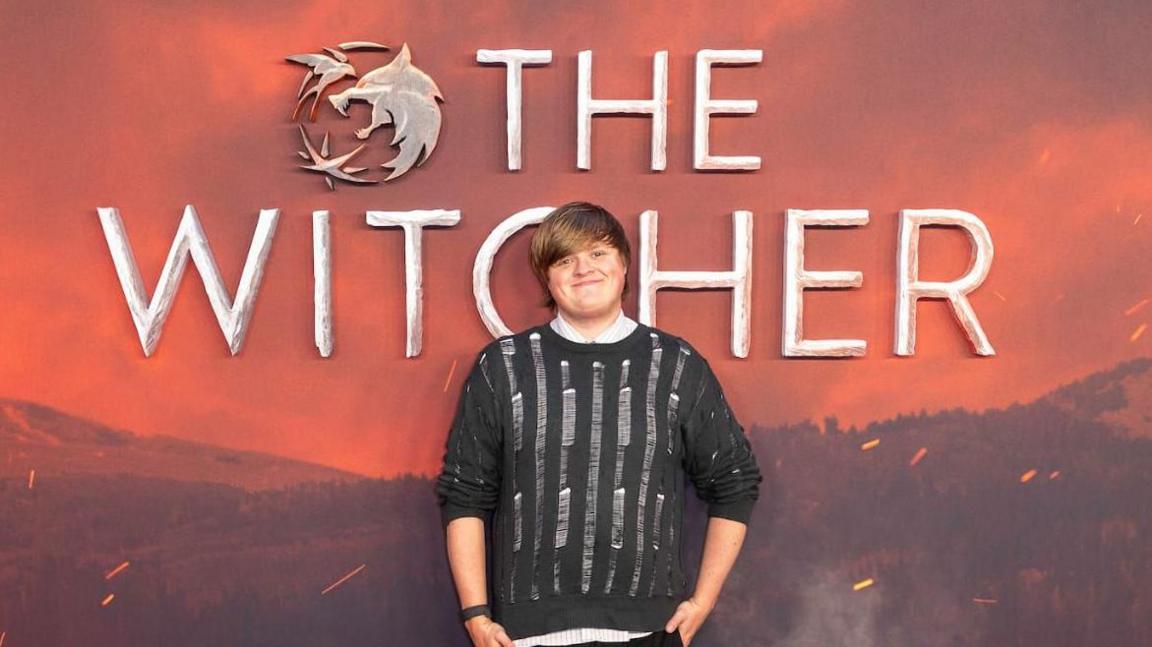 Dawson is wearing a white shirt and a black jumpter over it. He has his hands to his side and is smiling into the camera in front of The Witcher promo poster at the premiere.