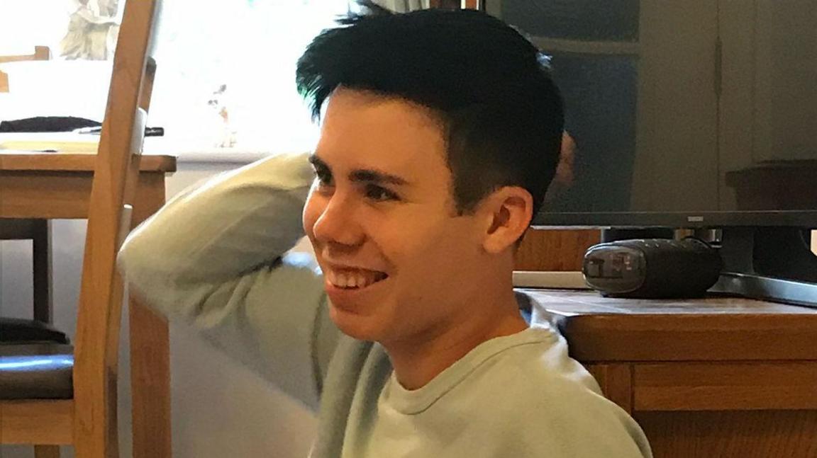 Alec Channing smiles in a room where he is sitting, possibly on the floor. He has black hair and wears a light-coloured top.