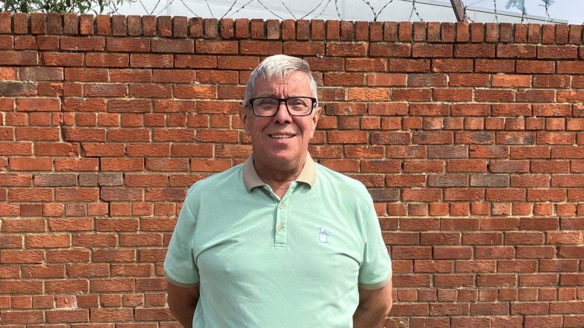 Alan Mardghum stares straight at the camera. He is stood in front of a red brick wall and has short grey hair and is wearing dark glasses, a green polo shirt and jeans