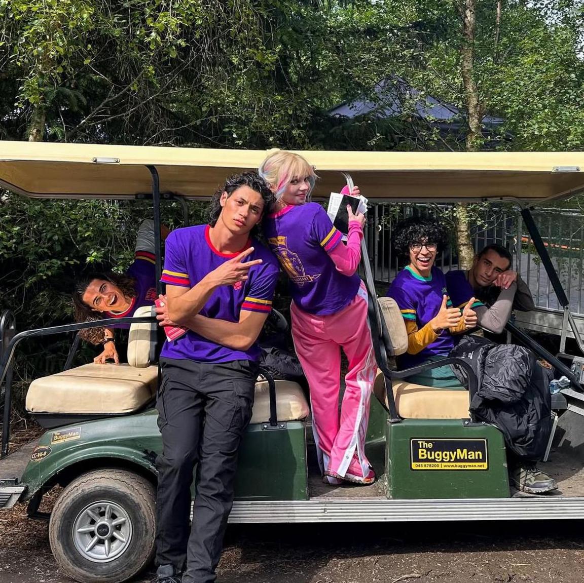 Cast of Wednesday either sitting on or standing next to a golf buggy. All wear distinctive purple tee-shirts. Actress Emma Myers has blonde hair and is wearing bright pink tracksuit bottoms and pink top under her tee-shirt.