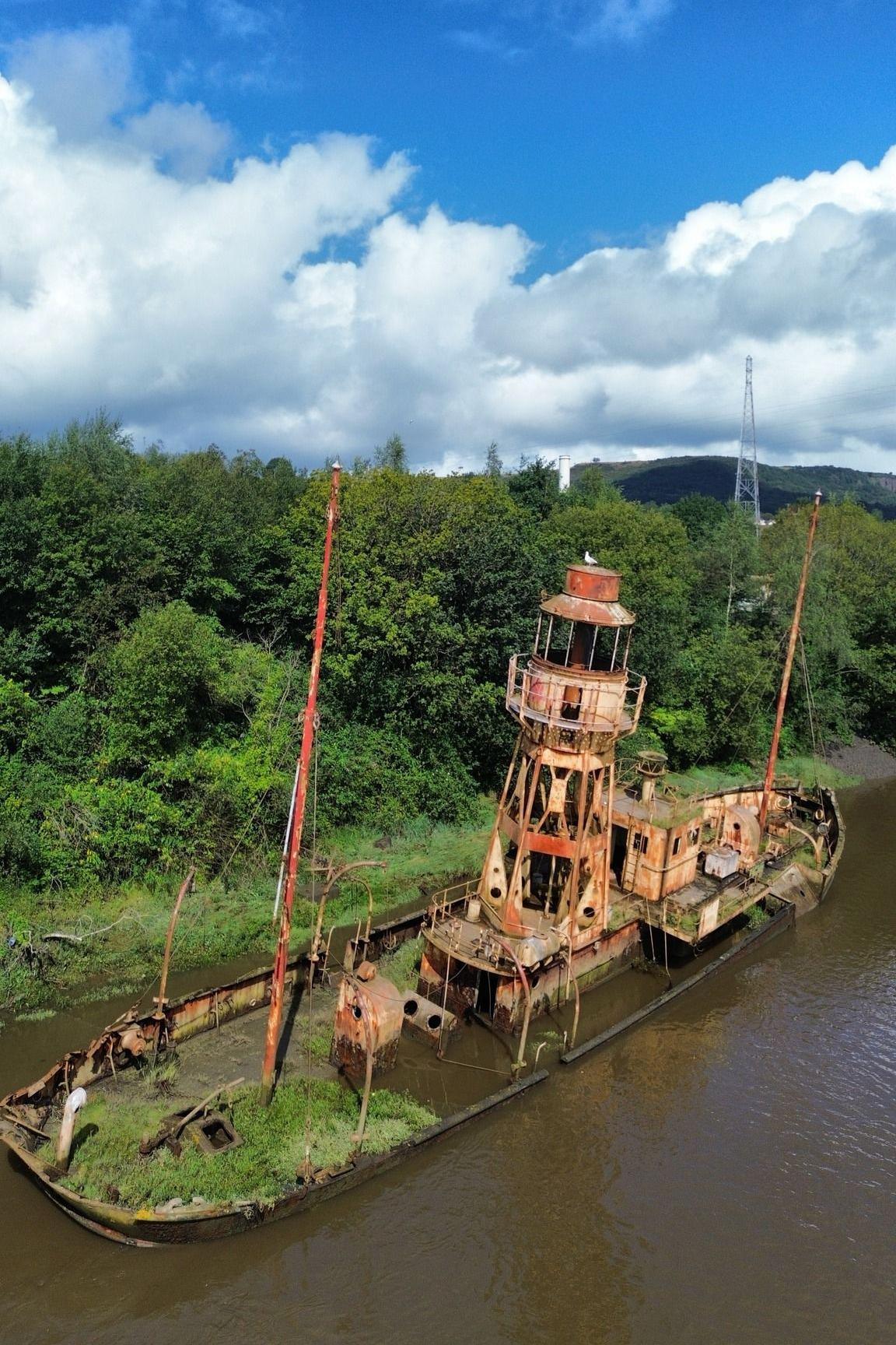 A rusty ship with a light house attached to it. It is slightly leaning, with greenery behind it and muddy water in front.