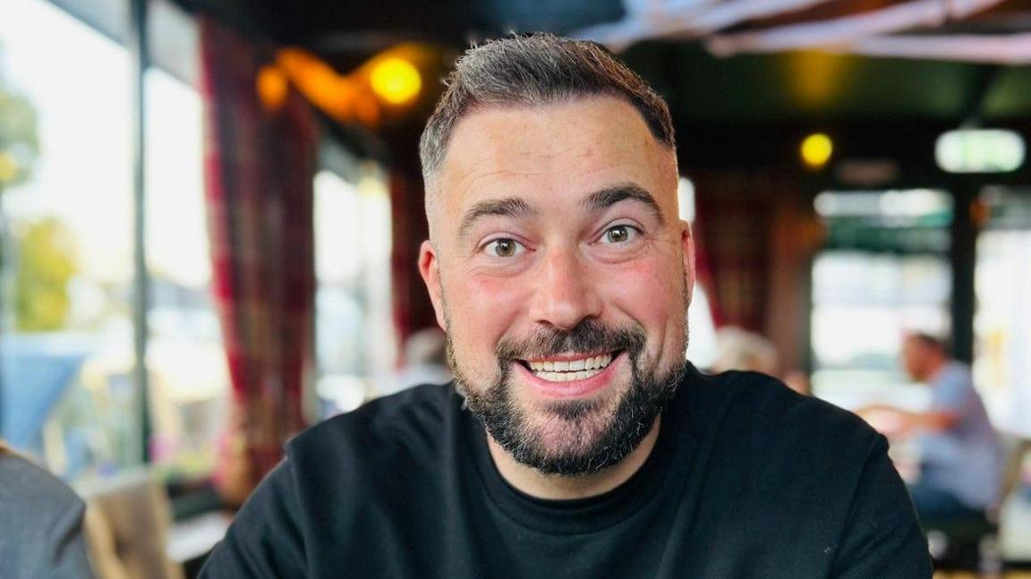 A man with short hair and a short, dark beard, wearing a black jumper.