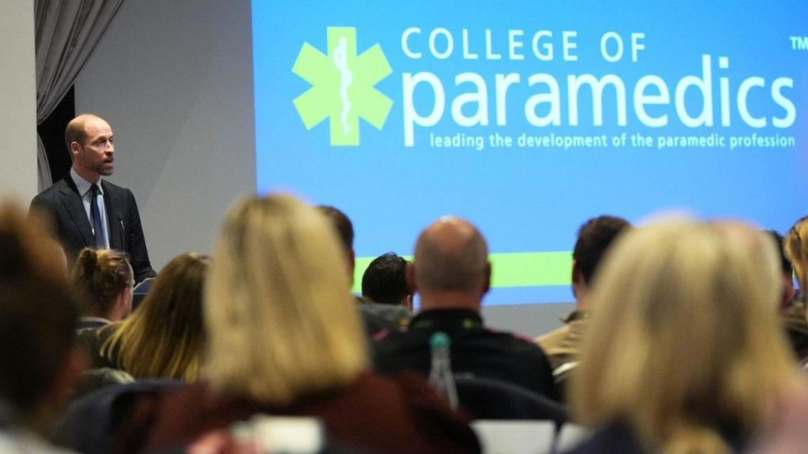 The Prince of Wales on the left of the screen speaks in front of an audience. Words including College of paramedics are on a blue screen at the front and the backs of heads of people in the audience are visible.