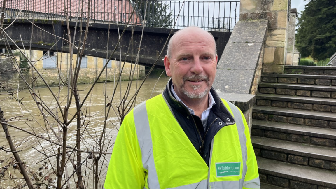 Nick Holder smiles at the camera. He is wearing a high vis jacket. Behind him is a stone bridge and a the river below it is high and brown. 