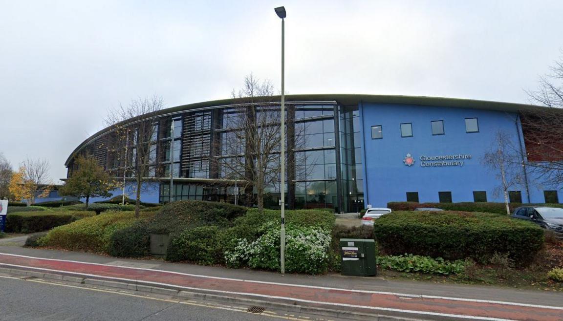 A screenshot from Google street view showing the Gloucestershire Constabulary headquarters. It is a large blue building with glass panels, situated behind trimmed hedges next to a road.