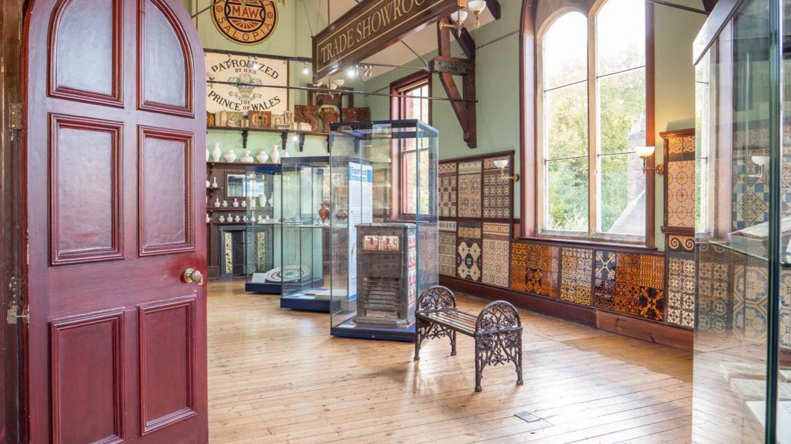 A room with a wooden floor and large windows with a red door and a number of glass display cabinets inside, and wrought iron benches.