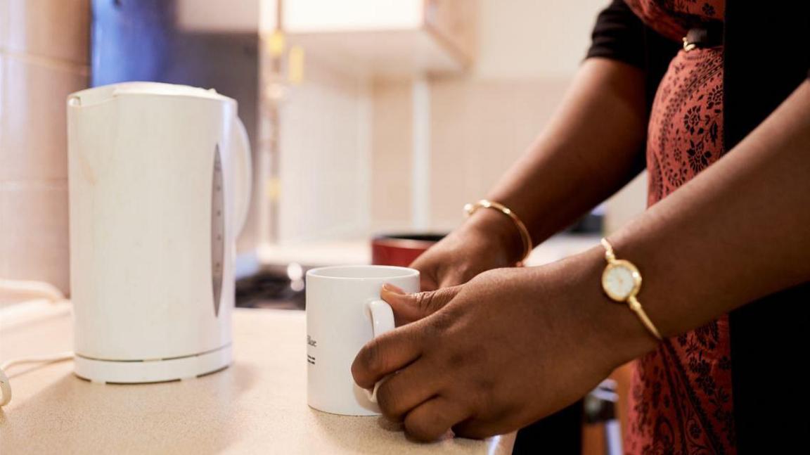 A woman wearing an orange dress stands by countertops in a kitchen. She is holding a white mug in front of white kettle.