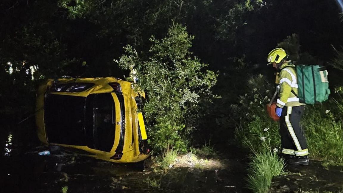 A yellow car can be seen on its side in the river, while an emergency worker in yellow high-viz is too the right with a green rucksack.