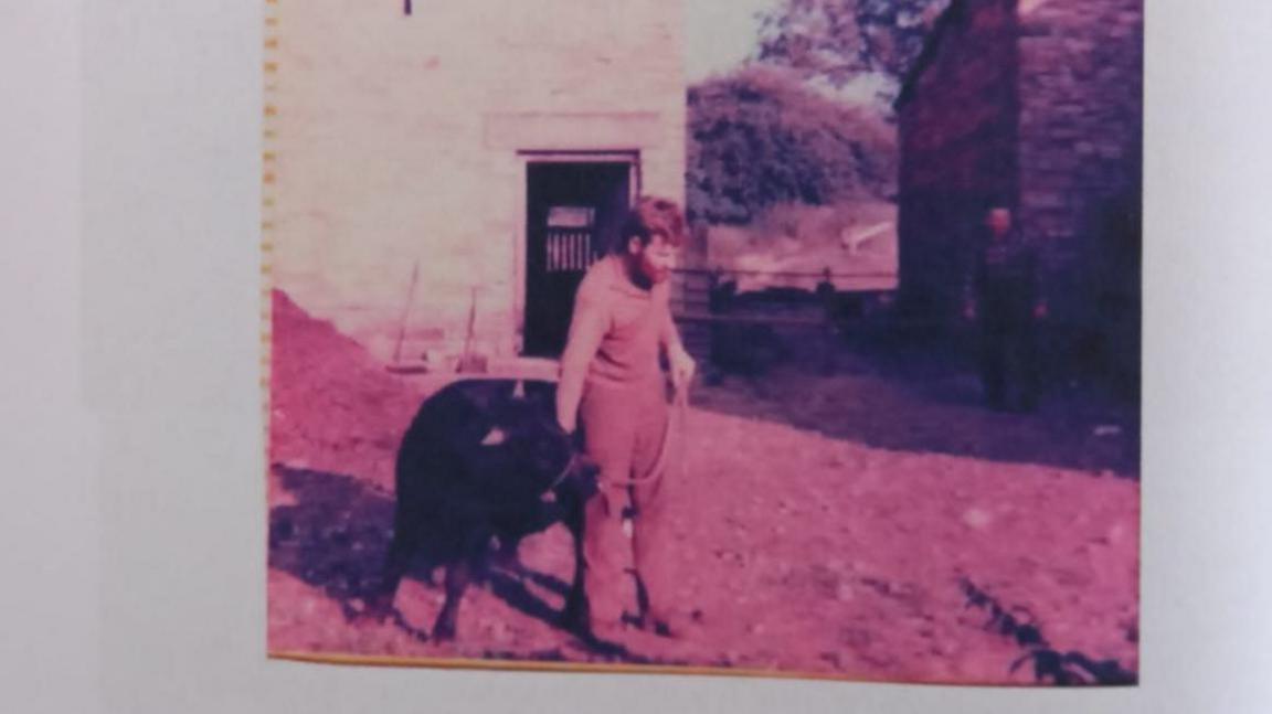 A photo showing a man, holding an animal in his right hand, in front of a stone building.