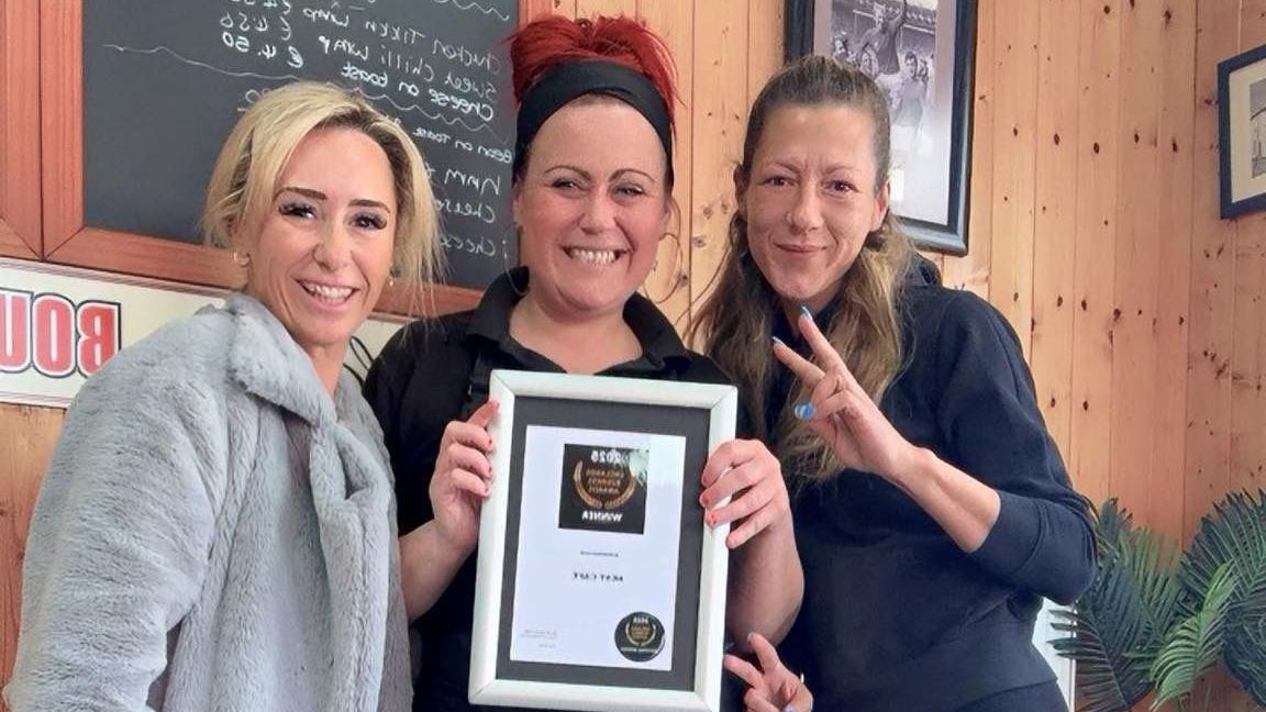 Three women are smiling in front of a wooden panelled wall, holding a certificate that says 'UK Small Business Awards winner'. The woman on the left has blonde hair tied back and is wearing a fluffly grey coat. The woman in the middle has tied back brown hair, is wearing a black shirt and apron and is holding a framed certificate. They woman on the right has long light brown hair and is wearing a navy blue top. She is holding two fingers up in a V sign for peace. THey are all smiling at the camera.