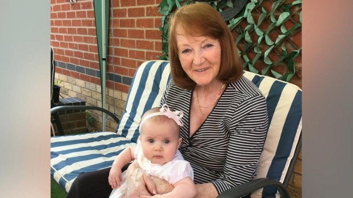 A woman in her 70s is sitting outside on a deckchair holding a baby. The baby is wearing a white and pink outfit with a bow in her hair.