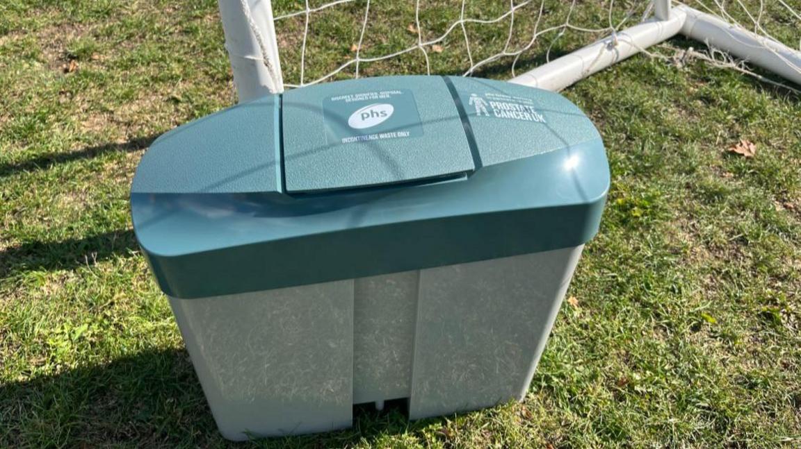 Image of a sanitary ware bin with a green top and a grey body.  The bin has been photographed on a football pitch in front of a goal net.