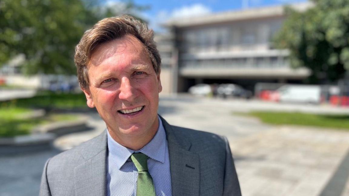 Angus Forbes wearing a patterned shirt, an olive green tie and a grey suit jacket in Armada Way in front of the Civic Centre.