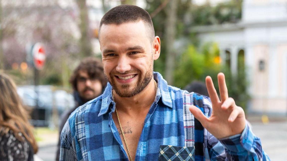 A man in a blue checked shirt, holding his hand up in a wave. He has brown hair cut in a shaved style and beard. He is in the street, with a stop sigh, trees and a white building just visible behind, as well as a man and a woman who are out of focus.
