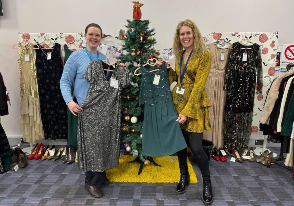 Siobhan Shaw with dark hair on the left wearing blue and Lizzie Dibble with blonde hair is on the right wearing yellow. They are both holding dresses, while other dresses and a Christmas tree are behind them.
