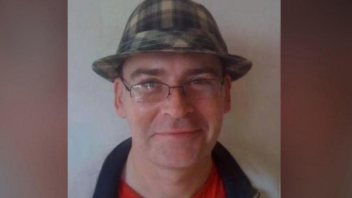 Christopher Corkill smiling. He's wearing glasses and a brown check tartan trilby hat.