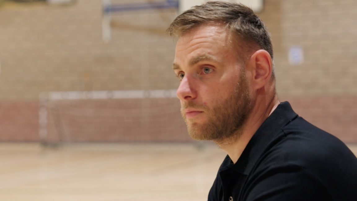 A head and shoulders profile view of a man with short straight brown hair and a beard, wearing a black top, inside a leisure centre, with brick walls and goal mouth in the background.