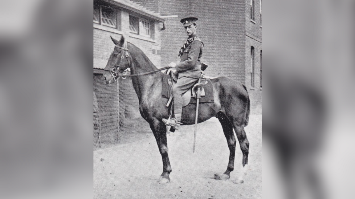 Black and white photo of Henry Bellamy in army uniform astride a large, dark horse. His rifle is strapped to the saddle. He is seen side-on from the camera and is looking into the lens, which is to his left. There are buildings behind him.