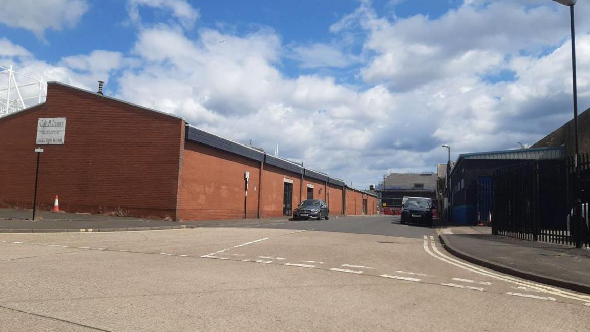 A row of red brick industrial units on Stobart Street. The picture is taken from the junction in the road. Two cars are parked outside the buildings.