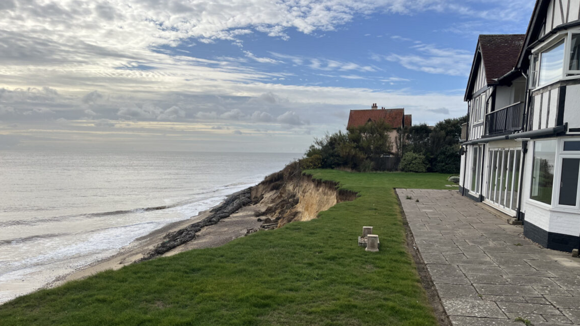 A heavily eroded coastline encroaches on a white house. The sea is on the left of the image and another house is in the distance.