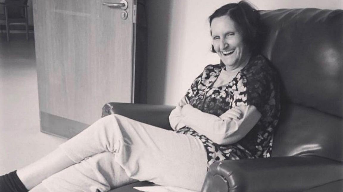 Minnie sits in a reclining chair, smiling warmly with her arms folded and legs stretched out. The photo is in black and white, taken in what looks like a hospital or care facility
