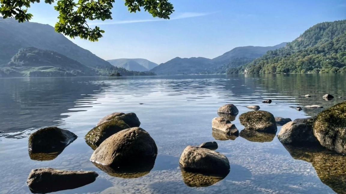 The waters of the lake are so still the rocks in the shallow end are reflected clearly. Beyond the lake, there are rolling green hills.