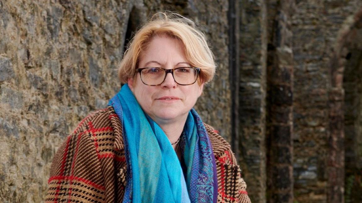 Prof Marion Gibson, sitting outside a historic stone building. She has fair, centre-parted hair and is wearing tortoiseshell glasses and a turquoise scarf over a thick red, black and brown checked coat.