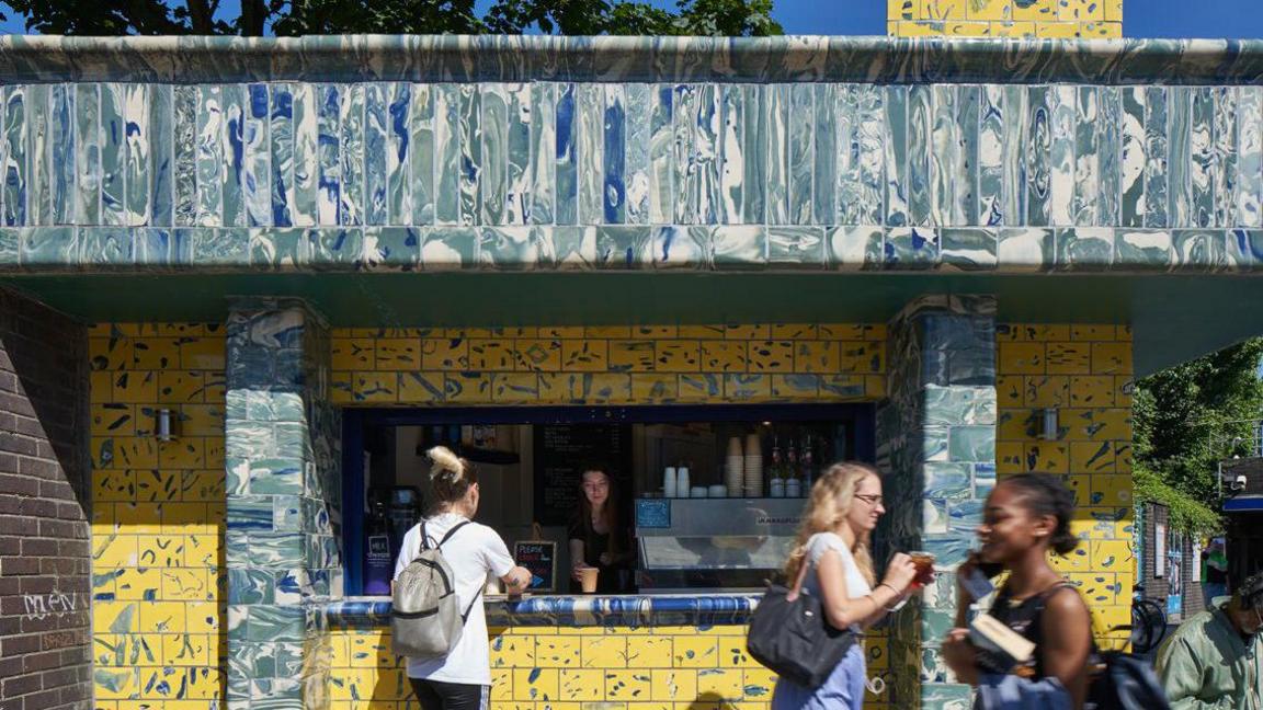 People walk past a coffee shop inside a commercial unit at a tube station. It is covered in yellow and marble effect tiles. A woman inside serves a coffee to a customer. 