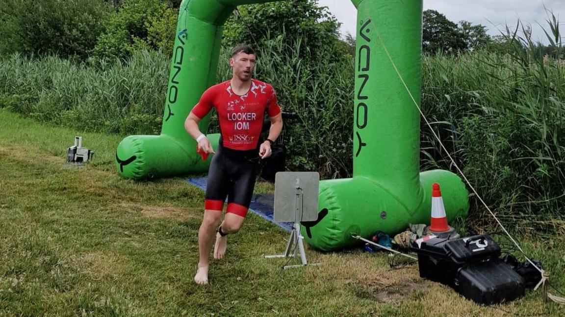 Matt Looker wears red and black lycra shorts and t-shirt, he runs barefoot past a large green inflatable arch on some grass.
