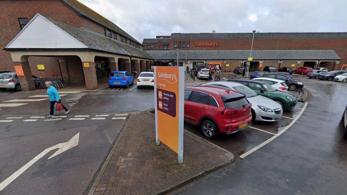 A Google Maps image of a car park in front of a Sainsbury's supermarket. There are dozens of cars parked and a handful of pedestrians walking. 