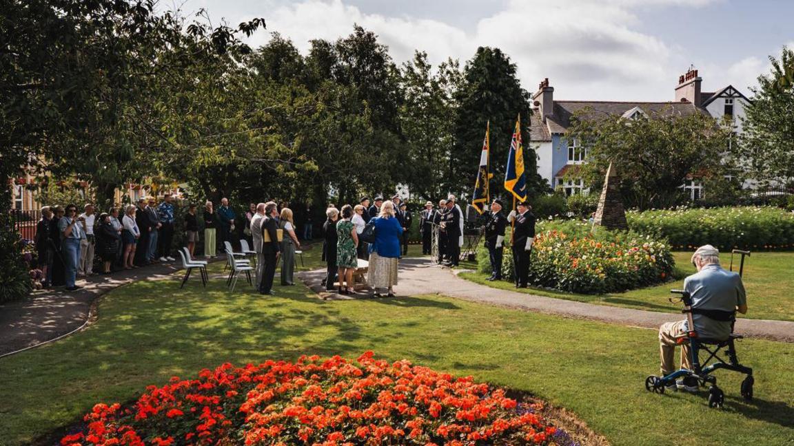 A wide photograph of about 40 people standing in a public garden: two people hold flags, a man stands by a lectern. In the foreground, to the right, an elderly man sits on a mobility aid, he wears a cap and holds a walking stick.