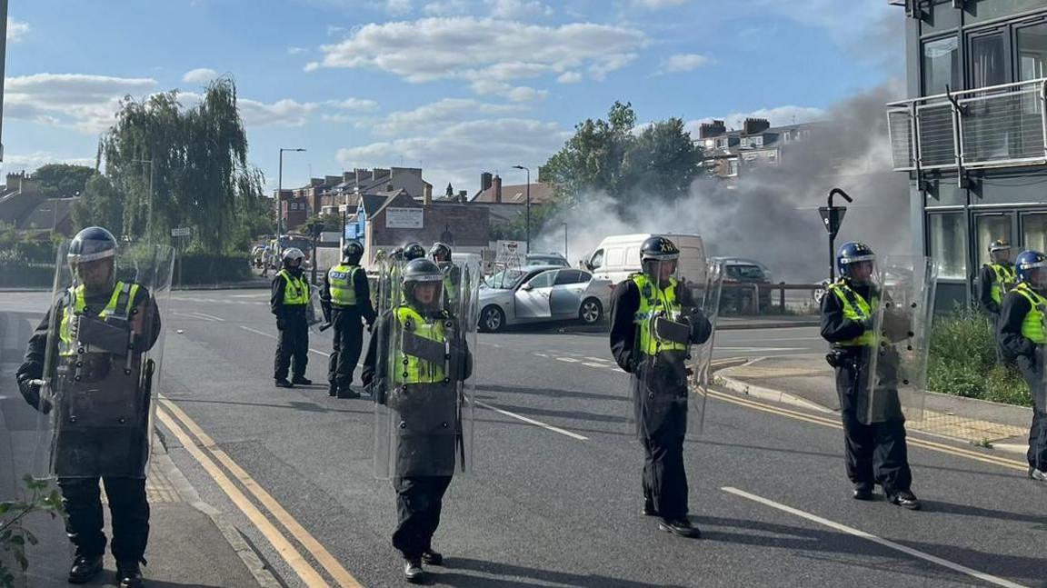 A row of riot officers lined across a street with burning cars in the far background