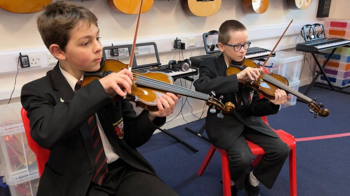 Two young boys wearing school uniforms with violins under their chins. They're sat in red chairs looking at sheet music. Behind them are three keyboards and guitars on the walls.