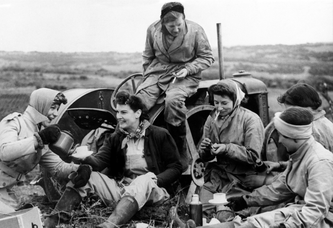 Image shows a group of young women in a field sitting by a tractor having tea out of flasks