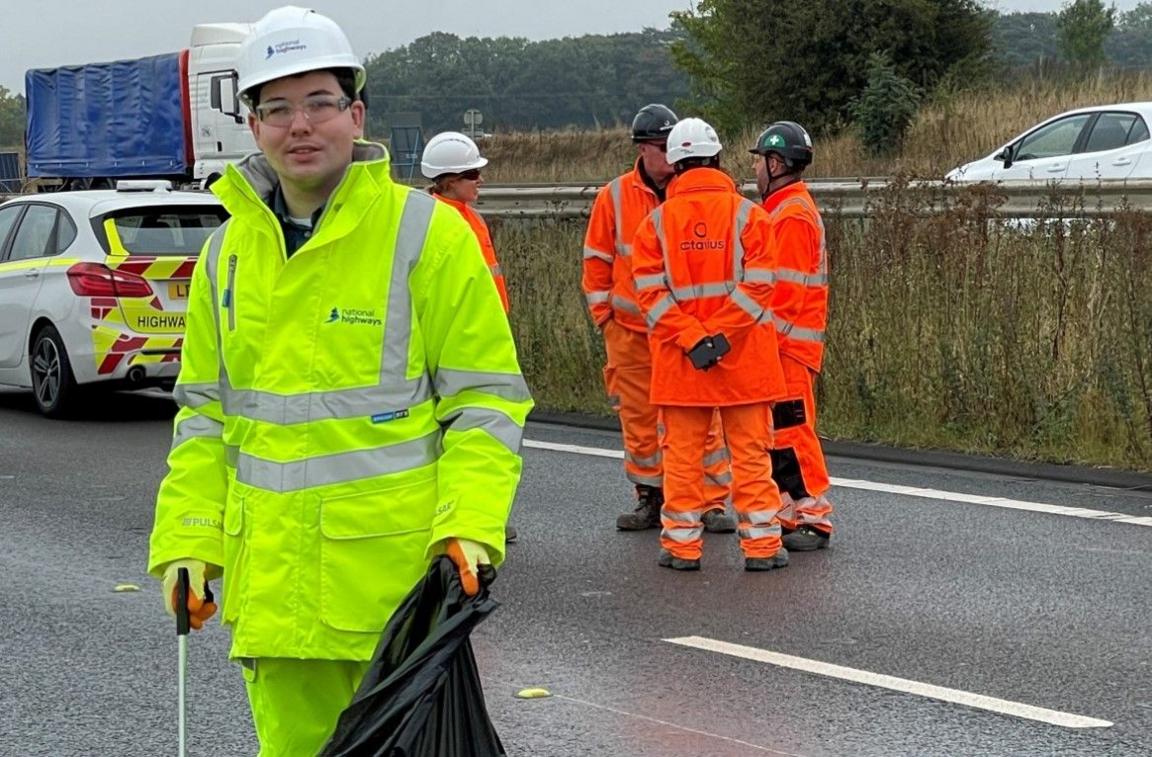 James Owen Thomas is carrying a black bag for collecting litter in on the closed part of the road, with other people in orange clothes and helmets behind him in another lane