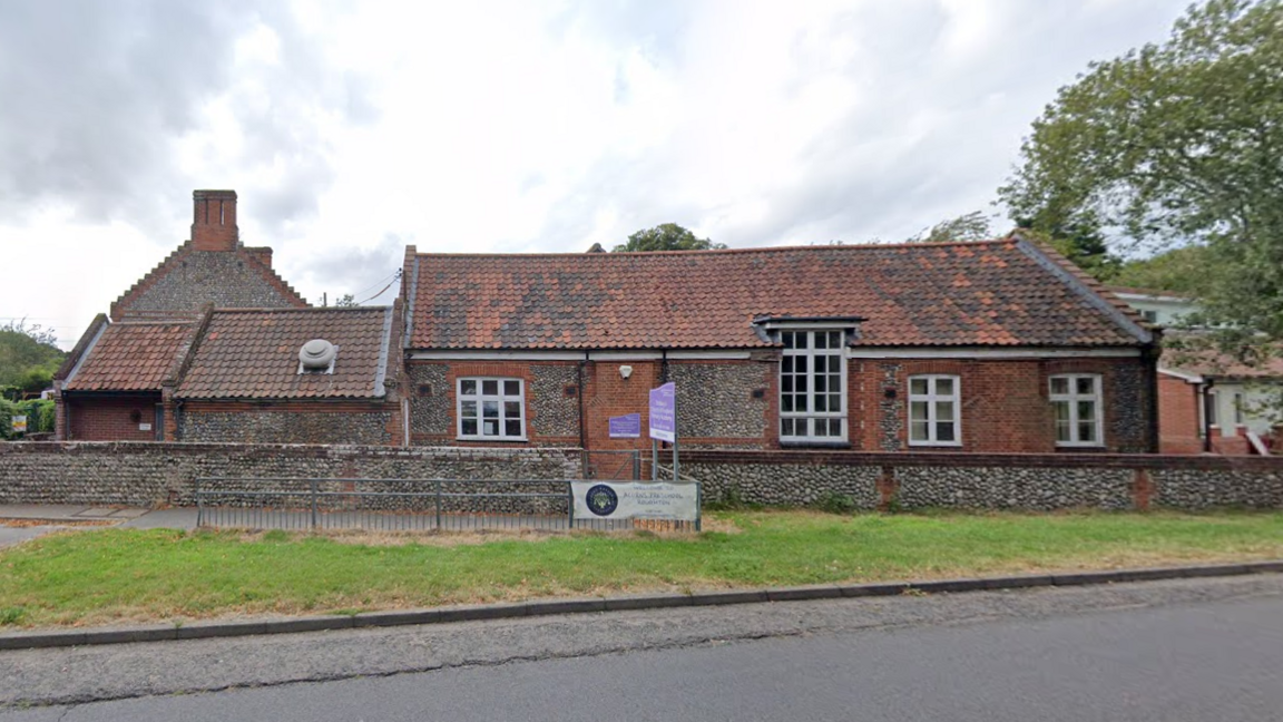 A red brick and stone building with a red tiled roof. The building has white window frames and a purple sign outside.