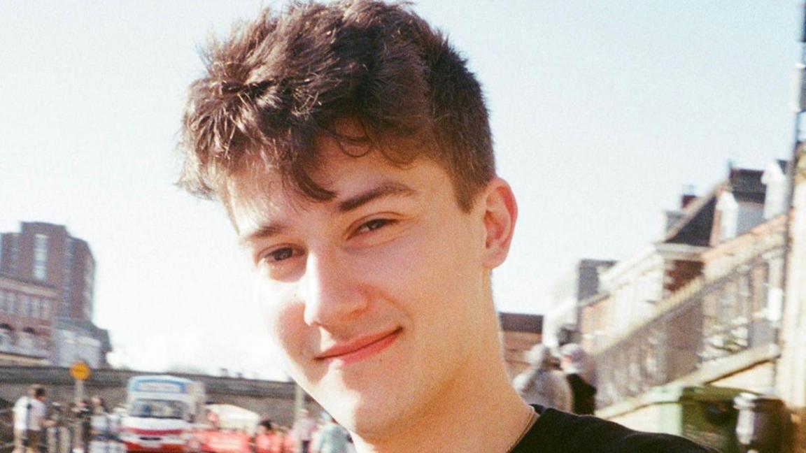 A smiling Ben Collins looking directly at the camera, standing outside with buildings in the background 