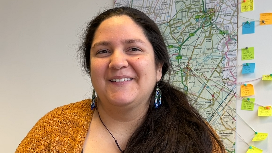 Ms Avilez is wearing a yellow knitted cardigan and long blue, white and green earrings. She has long brown hair, in a ponytail over her shoulder, brown eyes and is smiling at the camera. Behind her is a map on the wall with string and notes pointing to, and sharing comments, about the town of March. 