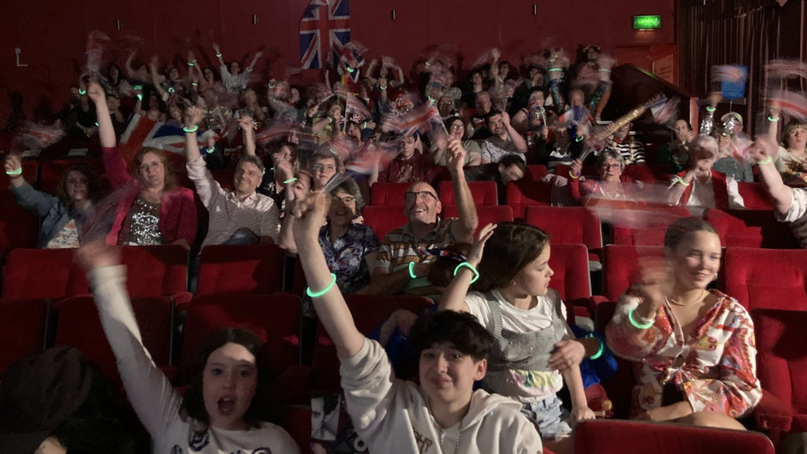 A large group of people are cheering and waving union flags as they sit in the cinema 