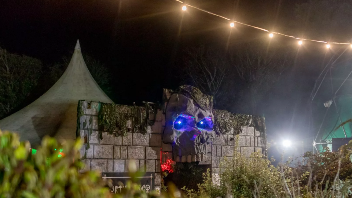 Festival entrance including a fake castle wall with a large skull with purple lights shining out of where its eyes would be over the entrance