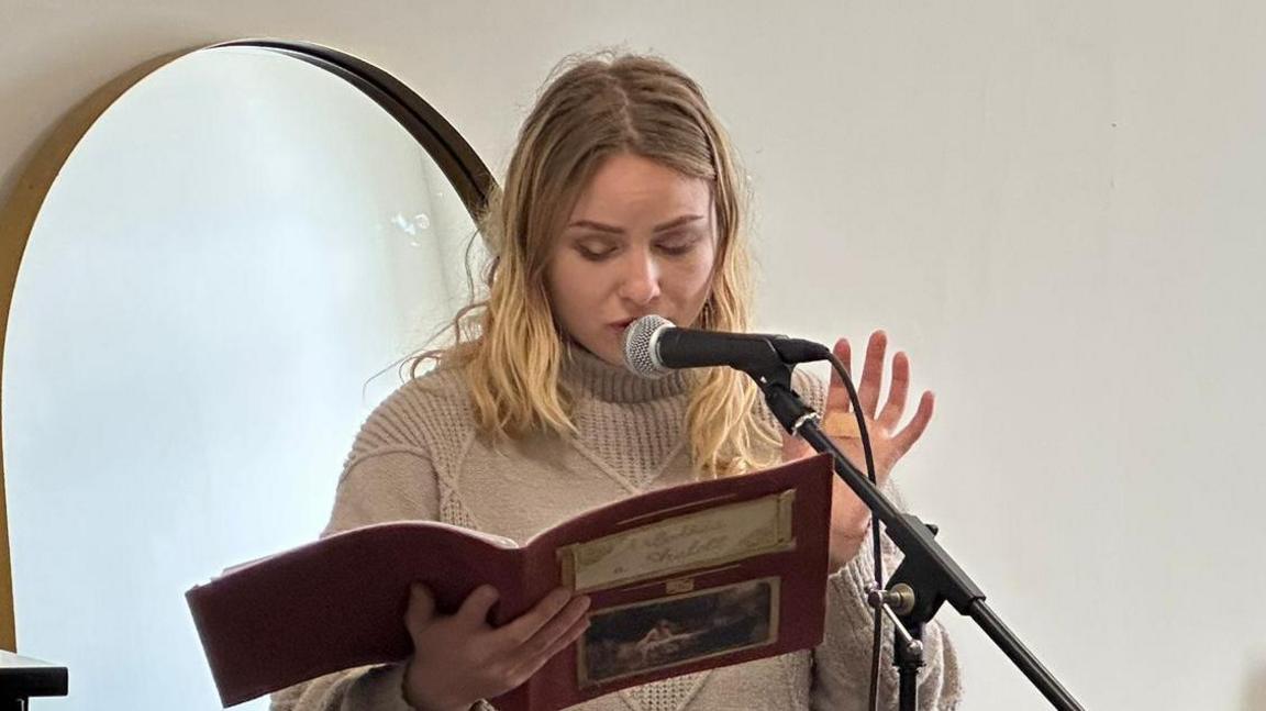 Elizabeth Freeborn singing into a microphone and holding a book while Elizabeth's head is down looking at it.