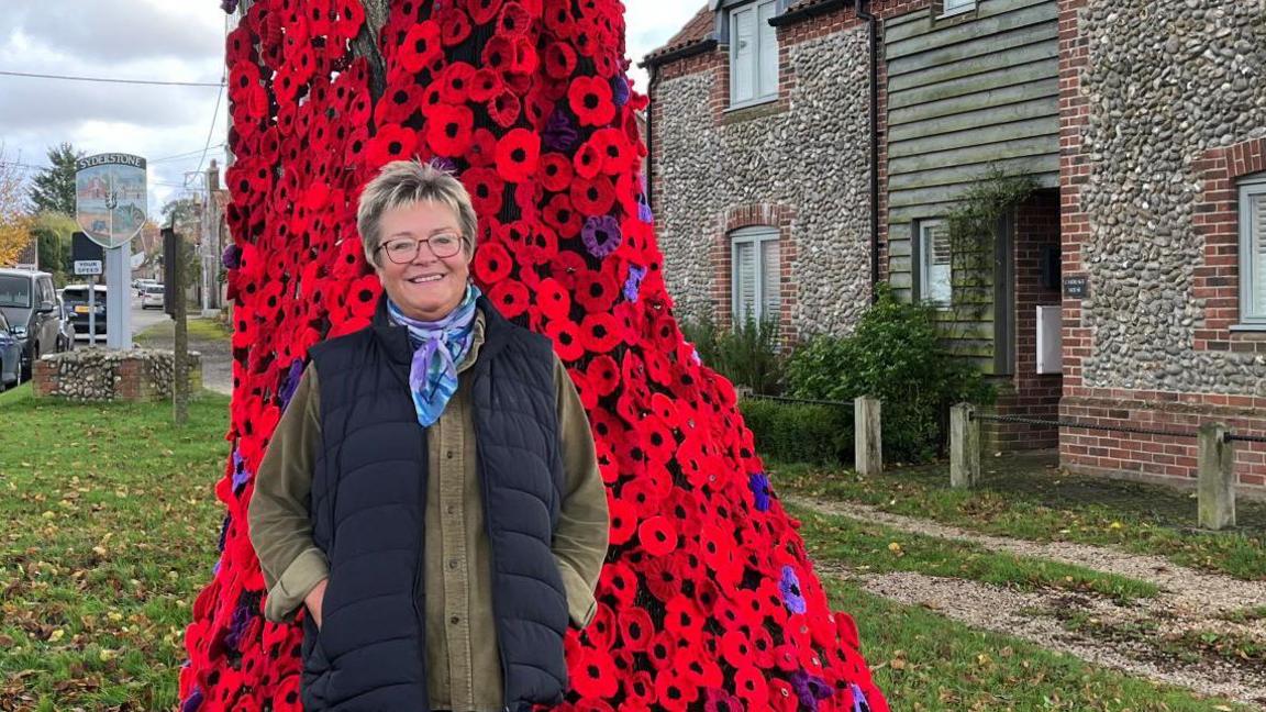 Woman stands in front of a tree which is covered in a cascade of knitted poppies in various shades of red and purple. The woman is smiling and wears a gilet and glasses.